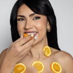 A woman smiles while holding a fresh orange slice and wearing more on her body, promoting natural beauty and nutrition.